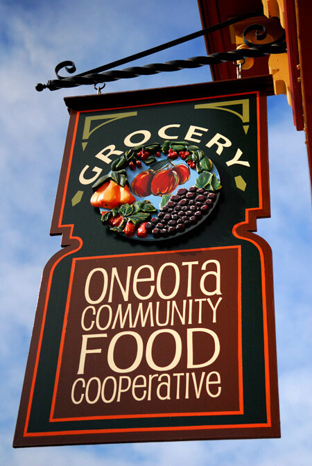 CBCCBE91-316F-43FA-BABC-413E77B97C1C Sign for Oneota Community Food Cooperative, featuring the word "Grocery" and a colorful depiction of fresh produce, hanging against a blue sky.