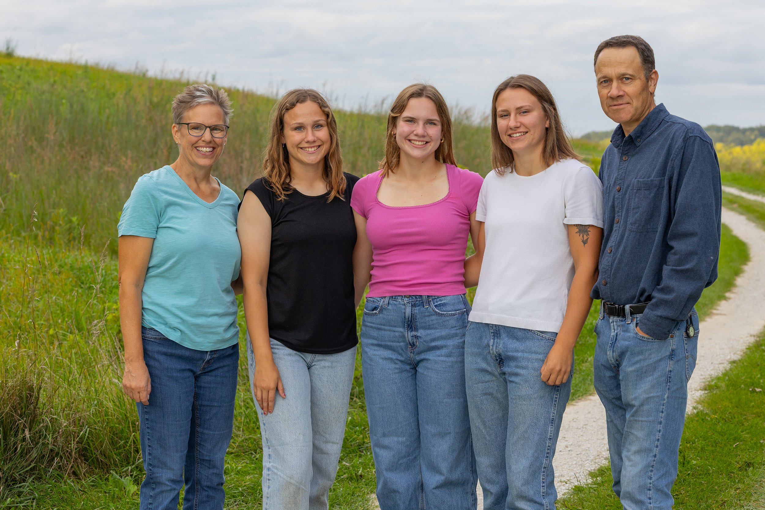 Copy+of+IMG_2871_screen Family portrait featuring five smiling individuals standing together on a grassy path, with a scenic landscape in the background. The group includes a woman in a light blue shirt, three young women in casual tops, and a man in a dark blue shirt, all dressed in denim.