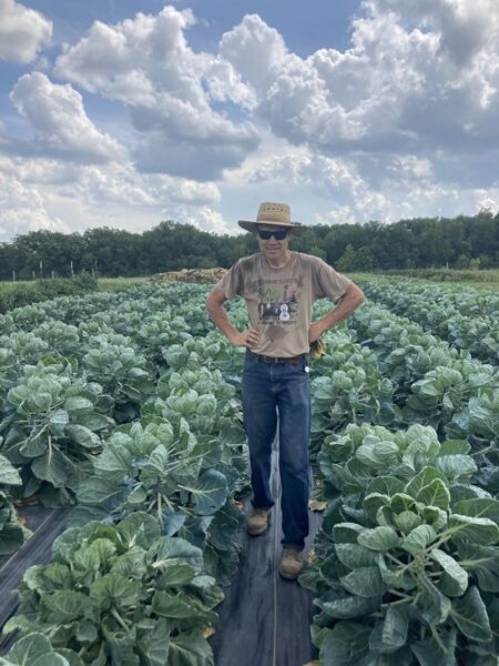 Farmer standing confidently in a field of vibrant green Brussels sprouts under a partly cloudy sky, showcasing sustainable agriculture practices.