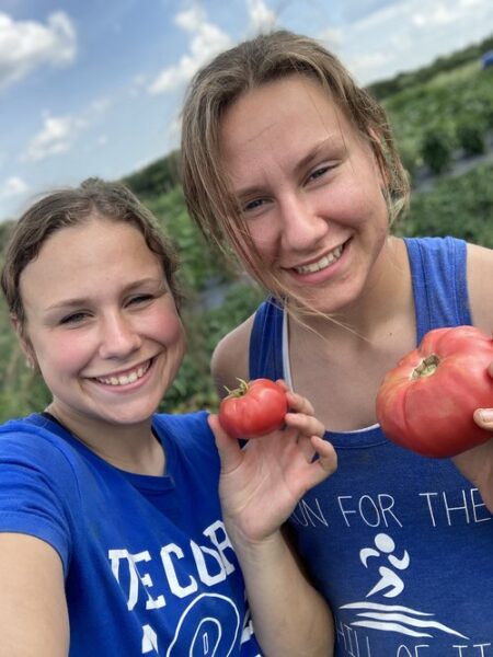 Two young women smiling and holding ripe tomatoes while standing in a lush green field. The image captures a joyful moment of harvesting fresh produce, highlighting the importance of farm-to-table practices and community gardening.