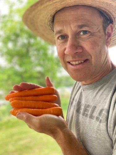 Man in a straw hat holding a handful of fresh, vibrant orange carrots outdoors, with greenery in the background.