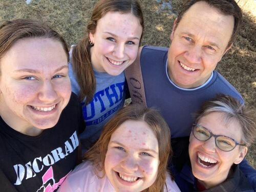 A happy family of five posing for a selfie outdoors, smiling and enjoying a sunny day. The group consists of two adults and three teenage girls, with diverse expressions and casual clothing.