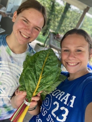 Two young women smiling and posing for a selfie while holding a large bunch of Swiss chard in a garden setting.